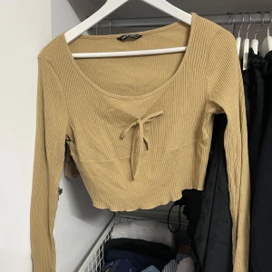 Beige ribbad topp från SHEIN - Säljer en superfin beige ribbad topp från SHEIN. Den har långa ärmar och en söt knytdetalj framtill. Toppen är croppad och perfekt för både vardag och fest. Den är i storlek L, passar M och i nyskick!