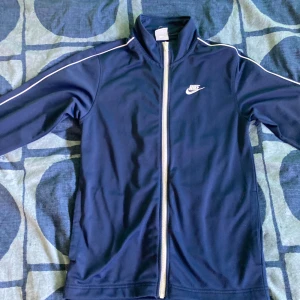 Mörkblå Nike träningsfleece  - Säljer en snygg mörkblå fleecetröja från Nike. Den har en vit dragkedja och vita detaljer längs ärmarna. Perfekt för träning & sport. Fleecen är i jättebra skick! Den har storlek XS men passar S