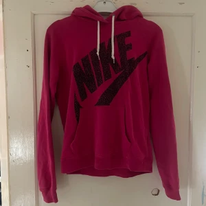 Rosa hoodie från Nike - Säljer en supercool rosa hoodie från Nike. Den har en stor Nike-logga i svart på framsidan och en mysig huva med vita snören. Perfekt för chilliga dagar eller när du vill ha en sportig look. Den är i bra skick och redo för nya äventyr!
