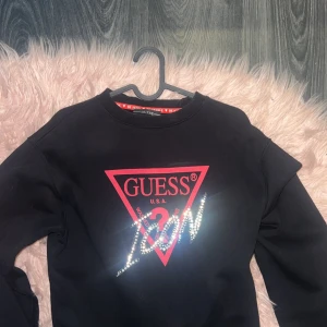 Svart tröja från Guess - Säljer en snygg svart tröja från Guess med det ikoniska röda triangellogotypen och texten 'ICON' i glittriga bokstäver. Perfekt för en casual look eller att klä upp med accessoarer. Tröjan har långa ärmar och är superbekväm.
