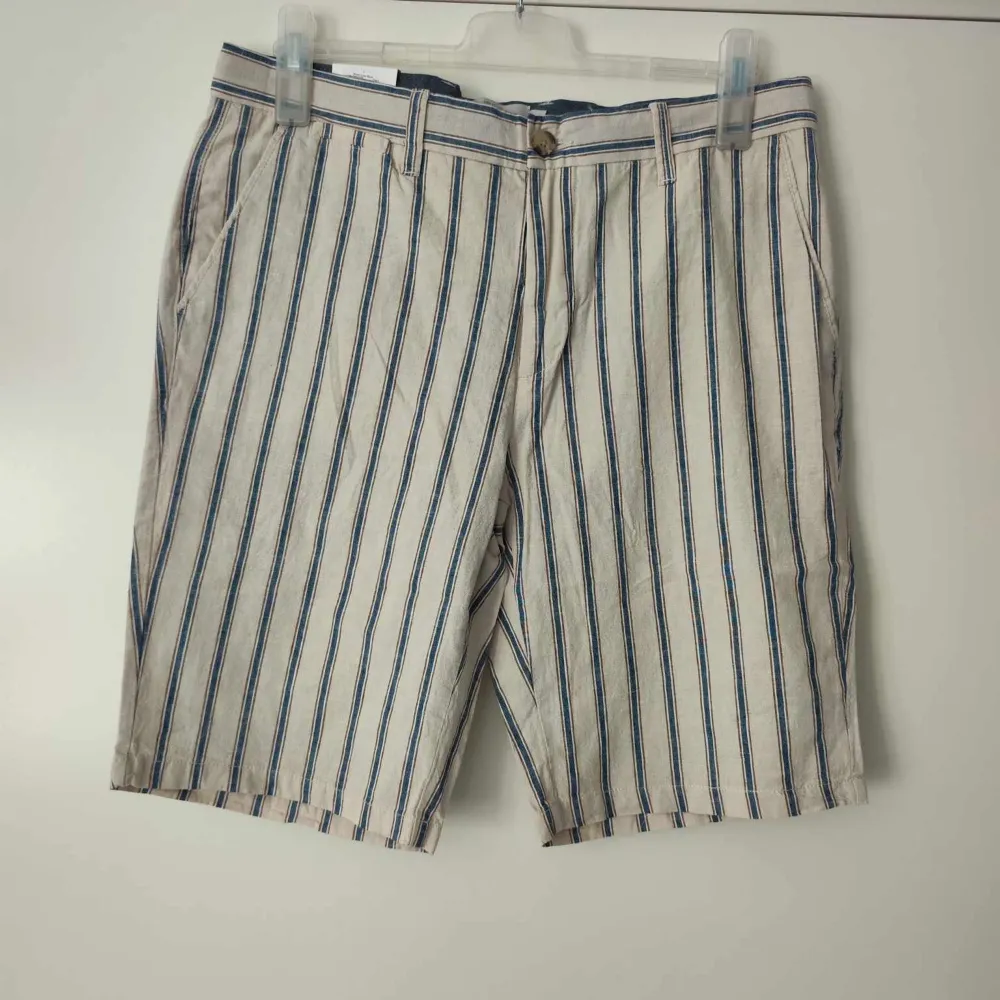 Säljer ett par snygga randiga shorts från Dressmann i storlek L. De är gjorda i en skön linneblandning, perfekt för varma sommardagar. Shortsen har ett klassiskt randigt mönster i blått och beige, med knappar både fram och bak. . Shorts.