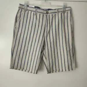 Säljer ett par snygga randiga shorts från Dressmann i storlek L. De är gjorda i en skön linneblandning, perfekt för varma sommardagar. Shortsen har ett klassiskt randigt mönster i blått och beige, med knappar både fram och bak. 