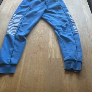 Blå mjukisbyxor från Hoodrich - Säljer ett par blå mjukisbyxor från Hoodrich i storlek S. Byxorna har en bekväm passform med resår i midjan och muddar vid bensluten. De har en cool vit text längs benen som ger en sportig look. Perfekta för chill eller träning!