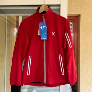 Röd träningsjacka från X TTL - Säljer en röd träningsjacka från X TTL, perfekt för  utomhusträning. Jackan har en svart panel på baksidan och vita detaljer runt dragkedjorna. Den är långärmad och har en dragkedja framtill. Använd som tredje lager. Nypris 800kr. Oanvänd 