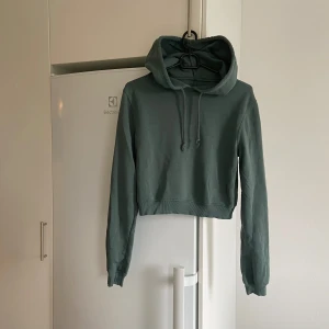 Grön croppad hoodie - Säljer en snygg olivgrön/khaki croppad hoodie med långa ärmar från Nelly. Den har en justerbar huva med dragsko. Otroligt skön och snygg samtidigt. Står ingen storlek men tippar på S. Den är i använt men bra skick.