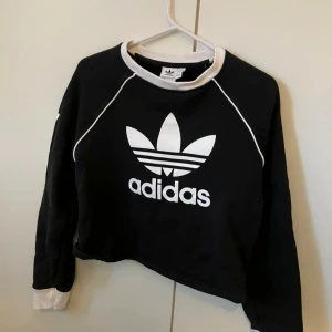 Svart och vit tröja från Adidas - Säljer en snygg svart tröja från Adidas med deras klassiska logga i vitt på framsidan. Tröjan har långa ärmar och vita detaljer vid kragen och ärmsluten. Perfekt för en sportig och avslappnad stil! storlek 38/M. Tröjan är en kortare modell. 