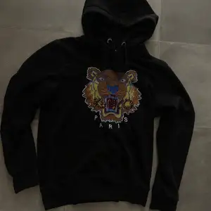 Kenzo-hoodie i mycket bra skick. Tröjan har en klassisk huva med dragsko och är tillverkad i ett mjukt och bekvämt material. Framtill finns ett stort, färgglatt broderi föreställande ett tigerhuvud, tillsammans med texten “KENZO PARIS” under tigern. 