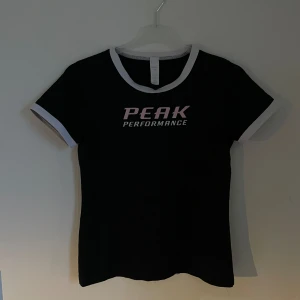 Svart t-shirt från Peak Performance - Säljer en snygg svart t-shirt från Peak Performance. Den har en vit kant runt halsen och ärmarna, vilket ger en cool kontrast. Perfekt för vardagsbruk eller träning. Texten 'PEAK PERFORMANCE' är tryckt i rosa och vitt på framsidan.