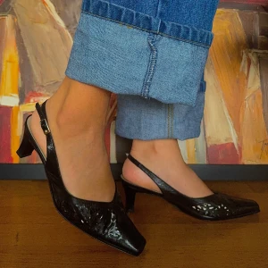 Svarta klackskor med slingback -  Carmen Lopez med slingback-design. Skorna har en spetsig tå och en elegant klack som ger en stilren look. Perfekta för festliga tillfällen eller en utekväll. De är gjorda i skinn och har en justerbar rem för bästa passform
