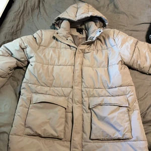 Beige pufferjacka med huva - Säljer en snygg beige pufferjacka med huva. Jackan är perfekt för kalla dagar och har en quiltad design som håller dig varm. Den har stora fickor framtill och stängs med dragkedja. Insidan är fodrad för extra komfort. Perfekt för vintern!