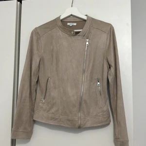 Beige mockajacka från Rosebud - Säljer en snygg beige mockajacka från Rosebud i storlek 36. Jackan har en cool asymmetrisk dragkedja och stilrena detaljer med knappar. Den är långärmad och perfekt för höst och vår. Jackan är i bra skick och passar både till vardags och fest.