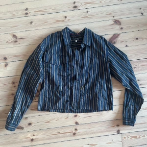 Randig skjorta från Armani Jeans - Säljer en snygg randig skjorta från Armani Jeans i storlek L. Skjortan har en cool mix av blå, beige och röda ränder och är gjord i ett lätt och bekvämt material. Den har långa ärmar och knappar framtill. Saknar knapp, rå kant nertill