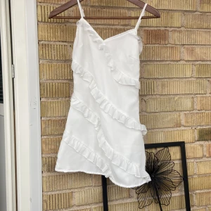 Vit klänning med volanger - Säljer en superfin vit klänning med volanger. Den är ärmlös och har smala axelband, perfekt för sommarens fester. Klänningen har en omlott-design som ger en snygg och luftig känsla. Materialet känns lätt och bekvämt, vilket gör den idealisk för varma dagar. Passar perfekt till både vardag och fest!