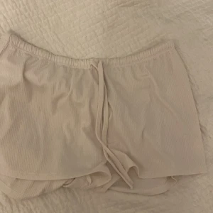 Beige shorts från Juicy Couture - Säljer ett par superbekväma och snygga beige shorts från Juicy Couture. De har en elastisk midja med knytband och är dekorerade med ordet 'Juicy' i glittriga stenar på baksidan. Perfekta för en avslappnad dag hemma eller en somrig utflykt!