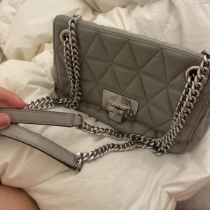 Grå quiltad axelväska från Michael Kors - Säljer en superfin grå axelväska från Michael Kors. Den är quiltad med ett snyggt mönster och har en kedja som axelrem. Väskan är i skinn och har en stilren design med en praktisk låsdetalj framtill. Perfekt för både vardag och fest!