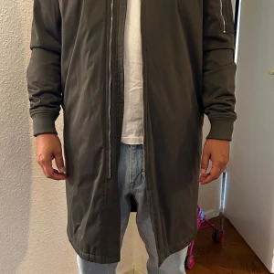 Grå lång jacka från Zara Man - Säljer en stilren grön lång jacka från Zara Man. Den har en dragkedja framtill och långa ärmar. Perfekt för höst och vår, med en modern och enkel design. Jackan är i bra skick och passar till många olika outfits. Killen på bilden är 1,70. 