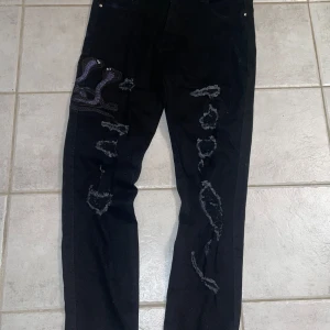 Svarta jeans med ormbroderi från Amiri - Säljer ett par svarta jeans från Amiri i bra skick. De har ett coolt ormbroderi i lila och svart på ena benet och slitna detaljer som ger en edgy look. Perfekta för en unik stil! 🐍