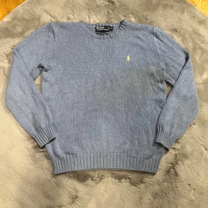  Stickad tröja från Ralph Lauren - Säljer en snygg ljusblå/grå stickad tröja från Polo Ralph Lauren i storlek M. Tröjan är i bomull och har en klassisk design med ribbade muddar och en gul broderad logga på bröstet. Perfekt för höst och vinter!