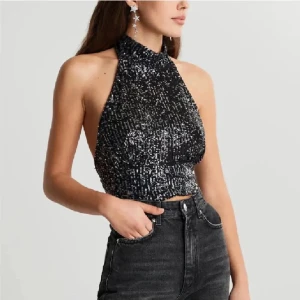 Svart glittrig halterneck topp - Snygg svart glittrig halterneck topp med öppen rygg och knytning. Perfekt för festliga tillfällen! Toppen är tight och ger en elegant look.