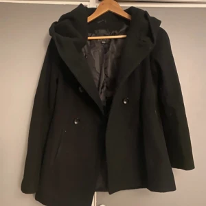 Svart kappa med huva - Säljer en stilren svart kappa med huva. Den har en klassisk design med knappar framtill och är perfekt för kyliga dagar. Jackan är i bra skick och passar både till vardags och lite finare tillfällen.