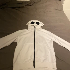 Vit c.p hoodie - Säljer en unik vit hoodie från C.P. Company med inbyggda glasögon i huvan. Perfekt för att sticka ut i mängden! Den har en dragkedja framtill och en stor ficka. Superbekväm och stilren design. Passar perfekt för höst och vår. Spricka i ett glas skriv för bättre bild