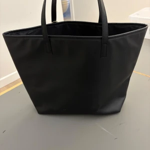 Svart tote bag från H&M - Säljer en stilren svart tote bag från H&M. Väskan är i syntetmaterial och har en enkel design med två handtag. Perfekt för vardagsbruk eller en dag på skolan/jobbet. Den är rymlig och lätt att matcha med olika outfits. 🥰🥰