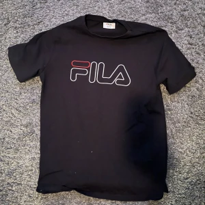 Svart t-shirt från FILA  -  Säljer en svart t-shirt från FILA i bra skick. Den har en stor logga på bröstet i vitt och rött. Perfekt för vardagsbruk eller träning. Passar till det mesta och är superbekväm! Använd nån gång men inget som syns. Storlek 158/164 (13-14 år)