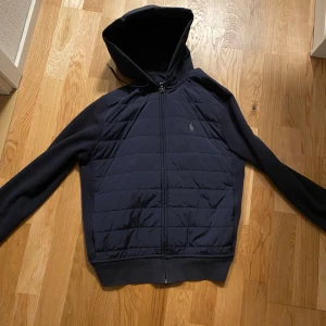 Mörkblå jacka med huva från Ralph Lauren - Säljer en snygg mörkblå jacka från Ralph Lauren. Den har en quiltad design på framsidan och en praktisk huva. Jackan är långärmad och har dragkedja framtill. Perfekt för höst och vår! NYPRIS ÄR 3500