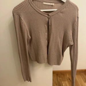 Beige ribbad kofta - Säljer en superfin beige ribbad kofta med knappar framtill. Den är perfekt för höst och vår, och har en mysig och stilren look. Koftan är långärmad och croppad, vilket gör den perfekt att matcha med höga jeans eller kjol. Perfekt för både vardag och lite finare tillfällen!