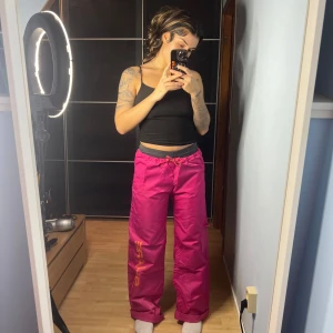 Rosa termo liknande byxor med tryck  - Säljer ett par coola rosa byxor med orange tryck på sidan. De har en loose passform och är perfekta för en avslappnad stil. Byxorna har snörning i midjan för justerbar passform. Perfekta för både vardag och fest! Passar XS-S