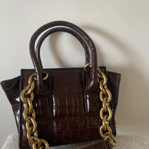 Brun krokodilpräglad handväska från Zara - Säljer en superfin brun handväska från Zara i krokodilpräglat skinn. Väskan har en cool kedjedetalj i guld och två handtag. Perfekt för att ge din outfit en lyxig touch! Kommer med en dustbag för förvaring.
