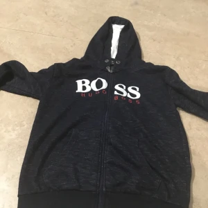 Svart hoodie från Boss - Säljer en snygg svart hoodie från Boss med vit och röd logga på bröstet. Den har en dragkedja framtill och en mysig huva med vitt foder. Perfekt för kyliga dagar eller som en stilren vardagströja. Passar både till jeans och joggers! Prisen kan diskutera.
