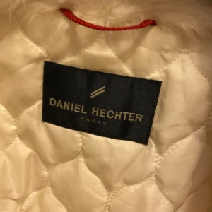 Vit jacka från Daniel Hechter - Säljer en elegant vit jacka från Daniel Hechter. Den har en quiltad insida och en mjuk pälsliknande krage. Perfekt för kyliga dagar när du vill hålla stilen. Jackan har knappar framtill och en ribbad nederkant för en snygg passform.