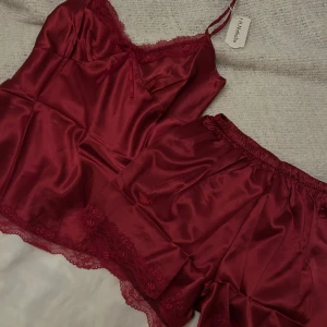 Vinröd satin pyjamas  - Säljer en helt ny superfin vinröd satin pyjamas. Setet består av ett linne med spetsdetaljer och en liten rosett framtill, samt matchande shorts med spetskant. Perfekt för mysiga kvällar hemma eller som en lyxig present. Storlek är dam S.
