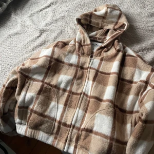 Rutig fleecejacka från Hollister - Mysig och varm fleecejacka från Hollister i beige och vit rutig design. Perfekt för kyliga dagar med sin bekväma passform och dragkedja framtill. Jackan har en huva och stora fickor för extra komfort. Perfekt för höst och vinter!