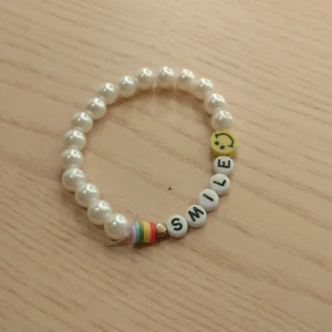 Pärlarmband med texten 'SMILE' - Superfint armband med vita pärlor och bokstavspärlor som bildar ordet 'SMILE'. Det har också en glad smiley och en regnbågsfärgad detalj. Perfekt för att sprida glädje och stil! 🥰