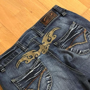 Blå jeans med broderad detalj - Säljer ett par snygga blå jeans med coola broderade detaljer på bakfickorna. Jeansen har en klassisk femficksdesign och är i bra skick!