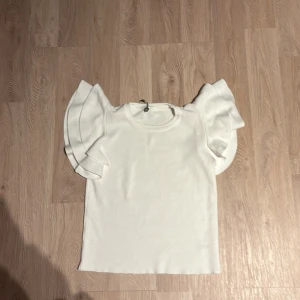 Vit topp med volangärmar - Säljer en söt vit topp med volangärmar. Perfekt för sommardagar eller en festlig kväll. Toppen har en rund hals och är i ett mjukt material som känns skönt mot huden.