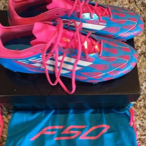 Adidas F50 fotbollsskor - Säljer ett par färgglada Adidas F50 fotbollsskor i blått och rosa. Skorna har en snygg mönstrad design med snörning och är perfekta för fotbollsträning. De är i mycket bra skick och kommer med en matchande skopåse.                   (Låda finns ej kvar)