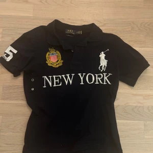 Svart kortärmad pikétröja från Ralph Lauren - Säljer en snygg mörkblå pikétröja från Ralph Lauren med broderad logga och texten 'NEW YORK' på framsidan. Tröjan har korta ärmar och nummerdetaljer på ärmen och ryggen. Perfekt för en Y2K stil tex! Säljes då den är för liten för mig som är 183.