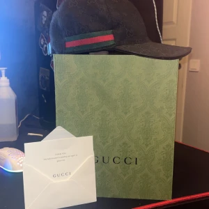 Svart keps från Gucci - Säljer en snygg svart keps från Gucci med deras ikoniska mönster. Kepsen har en grön och röd randdetalj på sidan och är i mycket bra skick. Perfekt för att ge din outfit en lyxig touch. Kommer med originalförpackning och dustbag. Kepsen är äkta och äkthets bevis finns. Kan ta emot byten så hör av er vid intresse. Möts i Göteborg / Kungsbacka 