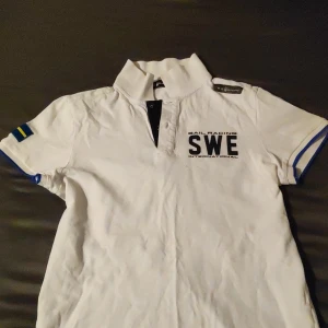 Vit pikétröja från Sail Racing - Säljer en snygg vit pikétröja från Sail Racing med texten 'SWE' tryckt på bröstet. Tröjan har korta ärmar med blå och gula detaljer samt en svensk flagga på ärmen. Perfekt för sommarens alla äventyr! 🌞