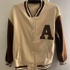 Säljer en snygg beige och brun bomberjacka med ett stort 'A' på bröstet. Jackan har långa ärmar och stängs med knappar. Perfekt för höst och vår, och ger en cool retrostil. Passar både till vardags och fest!