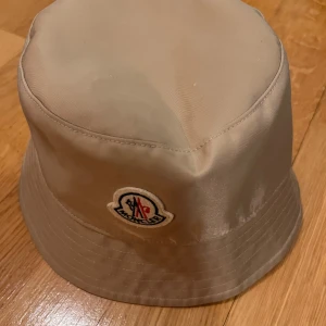 Beige bucket hat från Moncler - Snygg beige bucket hat från Moncler i mycket bra skick. Den har en broderad logga framtill och är perfekt för sommaren. Hatten är gjord i ett lätt och bekvämt material, vilket gör den idealisk för soliga dagar.