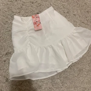 Vit kjol med volangdetaljer - Superfin vit kjol i ett mjukt och glansigt material. Den har en söt volangdetalj längst ner som ger en extra touch av stil. Perfekt för sommarens alla fester eller en dag på stan. Kjolen är kort och har en bekväm passform. Ett måste i garderoben för en chic look!