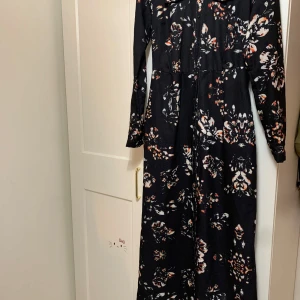 Blommig långklänning från Annah Hariri - Säljer en elegant långklänning från Annah Hariri med ett vackert blommönster i svart, vitt och orange. Klänningen har långa ärmar och en figursydd passform som ger en smickrande siluett. Perfekt för höst- och vinterfester!