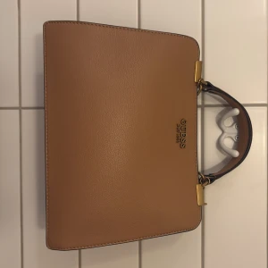 Beige handväska från Guess - Säljer en stilren beige handväska från Guess i mycket bra skick. Väskan har en elegant design med guldfärgade detaljer och en praktisk dragkedja. Perfekt för både vardag och fest! 👜✨