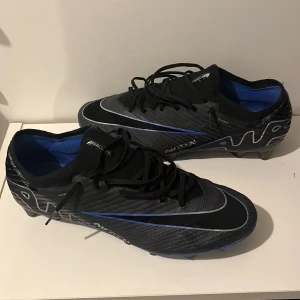 Svarta Nike fotbollsskor med blå detaljer - Säljer ett par svarta Nike fotbollsskor i mycket bra skick. De har snygga blå detaljer och snörning för perfekt passform. Skorna har en modern design med Zoom-teknologi för extra komfort och grepp på planen. Perfekta för fotbollsspelare som vill ha stil och funktionalitet.