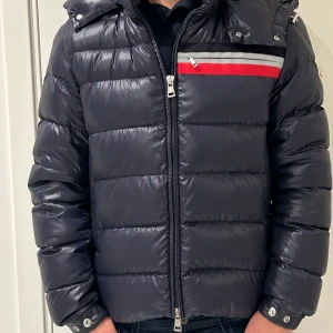 Svart pufferjacka från Moncler - Säljer en snygg blå pufferjacka från Moncler. Den har en cool röd och grå randdetalj på bröstet och en praktisk huva. Jackan är perfekt för kalla vinterdagar och har en dragkedja framtill. Den är i mycket bra skick och redo för nya äventyr!     -nypris 16000