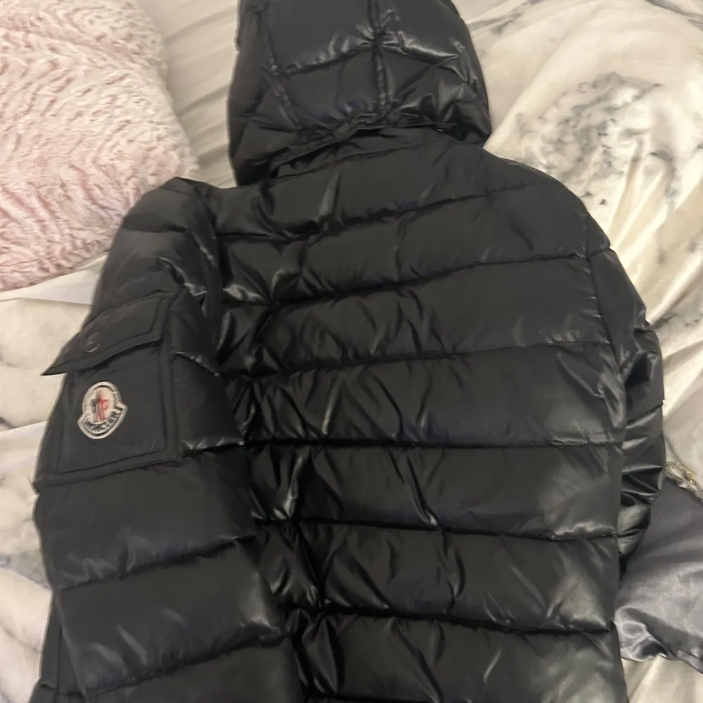 Säljer en svart dunjacka från Moncler. Den är varm och perfekt för vintern med en snygg glansig finish. Jackan har en dragkedja och en logga på ärmen. Det finns en liten skada vid ärmslutet som behöver fixas. Perfekt för dig som vill ha stil och komfort i kylan!. Jackor.